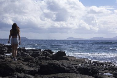 Volkanik kayalar ve mavi okyanusta yaz kıyafetleri giyen kadınlar.