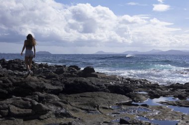 Volkanik kayalar ve mavi okyanusta yaz kıyafetleri giyen kadınlar.