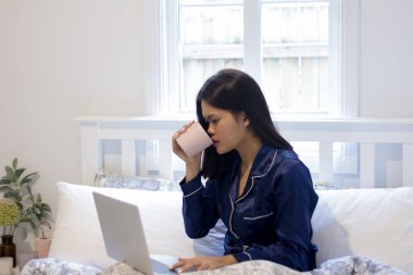 Genç Asyalı gülümseyen kadın elinde pembe bir kupayla dizüstü bilgisayarda çalışıyor. Mutlu sabahlar