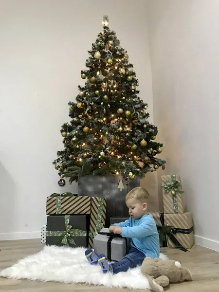Bir çocuk Noel süslenmiş bir ağacın yanında oturur ve hediyesini açar.