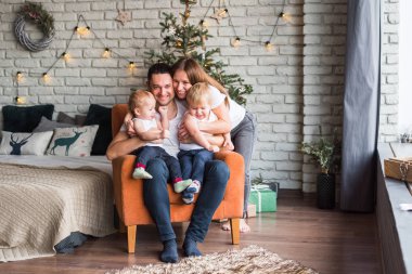 Anne, baba ve iki oğul Noel zamanı turuncu bir sandalyede oturup birbirlerine sarılırlar ve gülümserler.