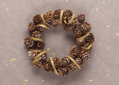 Doğal çam kozalaklarından yapılmış Noel çelengi ve bej kağıt arka planda altın kurdeleler.