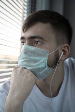 Gün batımında tıbbi maskeli genç bir adam pencereden dışarı bakar. Kulaklıklarla. Kişisel tecrit.
