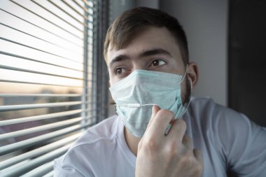 Tıbbi maskeli genç bir adam pencerede oturur ve kulaklıkla müzik dinler. Karantina.