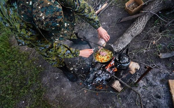 A man in a hunting uniform is cooking at the stake. Cauldron with soup. Bonfire in the forest