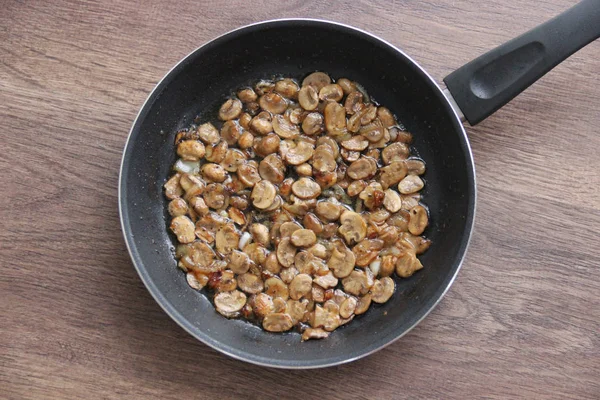 Fried small mushrooms in oil in a pan on a wooden table, top view