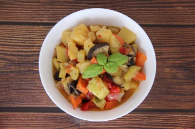 Vegetable stew with a Basil leaf in a white bowl on a wooden table, top view