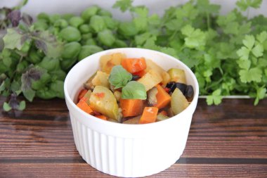 Tahta bir masada, beyaz bir kupada sebze güveci taze yeşilliklerin arka planına karşı.