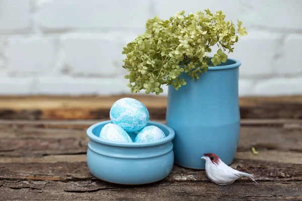 Decorative dyed easter eggs in blue pot with green hydrangea in skyey ceramic vasa and a little bird on brown wooden background with copy space