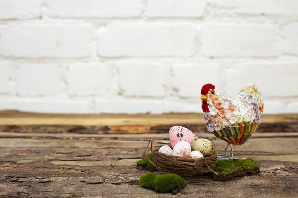 Украшение пасхального стола разноцветными яйцами в гнезде, корзина с декоративной курицей в цветах и мхом на деревянном столе и белом кирпичном фоне. Принято. Место для текста — стоковое фото