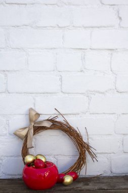 Paskalya altınları ve kırmızı yumurtalar kabın içinde, ahşap masa üzerinde beyaz tuğla arka plan yakınında el yapımı çelenkler var. Dikey, boşluğu kopyala.