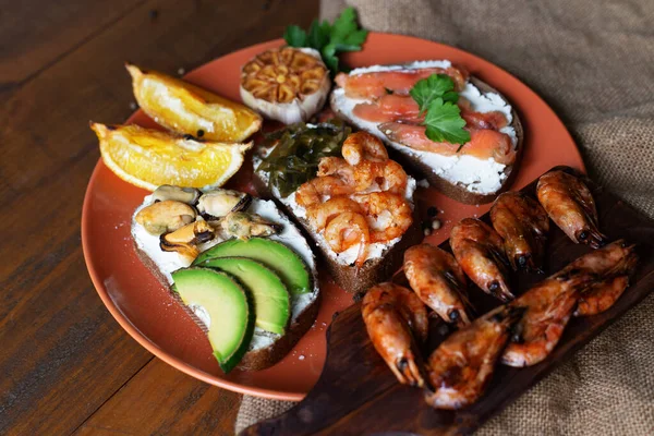 Seacuterie board with grilled shrimps, sandwiches with seafood, grilled garlic and lemon on plate on wooden background with sackcloth. Sandwiches with shrimps, avocado, red fish, mussels and sea kale.