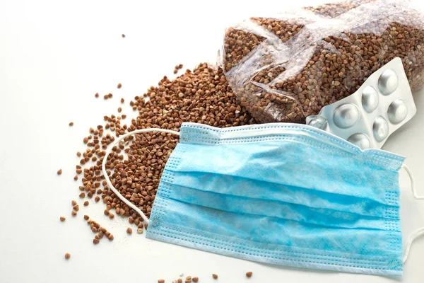 Coronavirus COVID 19 salgını sırasında Groats, masalar ve tıbbi cerrahi maske tedarik ediliyor. Beyaz arka planda karabuğday. Karantina ve sosyal uzaklık kavramı