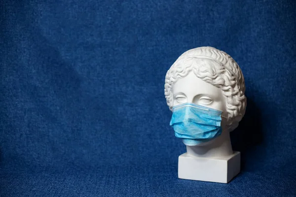 Yüzünü kaplayan tıbbi maskeli bir tanrıça büstünün beyaz alçı heykeli. Coronavirus COVID-19 konsepti. Kopya alanı olan klasik mavi arkaplan. Yatay