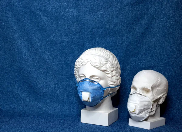 White plaster statues of a bust of goddess and a skull in a medical surgical masks covering their face. Coronavirus COVID-19 concept on classic blue background with copy space. Horizontal