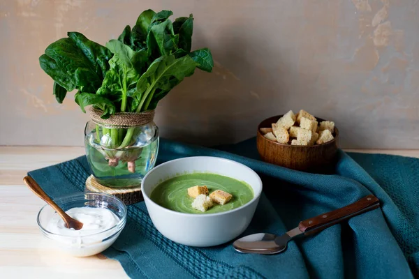 Healthy green soup puree in a grey bowl with crouton, raw spinach leaves in glass jar and sour cream with spoon on green tablecloth on wooden board