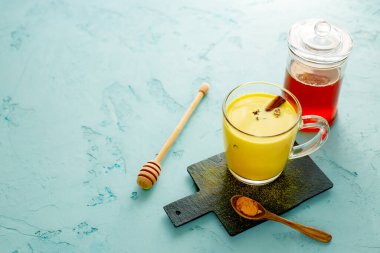 Seramik tahtasında tahta kaşık olan zerdeçal sarısı sütlü latte. Cam kavanoz bal. Mavi arka planda Hindistan içeceği ve tarçınlı cam bardak. Detoks sağlıklı, sıcak bir içecek. Boşluğu kopyala