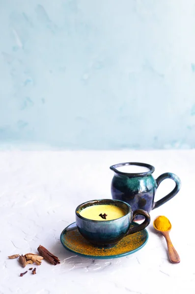 Baharatlı zerdeçal altın sütlü latte. Seramik sürahi süt, ahşap kaşık zerdeçal, tarçın çubukları, karanfil ve kakule. Açık mavi arka planda sağlıklı bir içecek. Uzayı kopyala, dikey