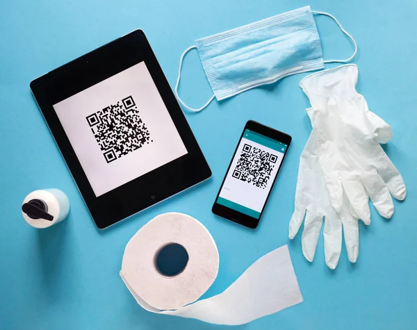 Coronavirus karantinası sırasında QR kodu taranıyor. Akıllı telefon, lateks eldiven, tuvalet kağıdı, sabun, dezenfektan, cerrahi maske ve mavi arka planda tablet bilgisayar. COVID-19 kontrol. Üst görünüm kavramı