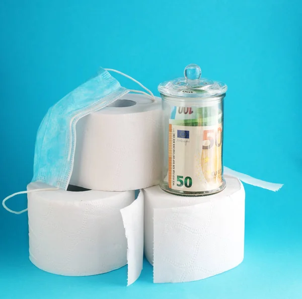 Tuvalet kağıdı, cam kavanozdaki para banknotları ve parlak mavi arka planda cerrahi maske. Coronavirus covid-19 için önemli şeyler kavramı.