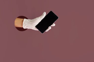 COVID-19 koruma konsepti. Tıbbi koruyucu eldiveni marsala renkli kağıdın arka planına karşı cep telefonu tutarak kağıt delikten uzat..