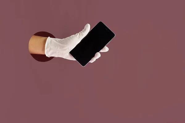 COVID-19 protection concept. Hand in medical surgical protective glove through paper hole holding mobile phone against marsala colour paper background.