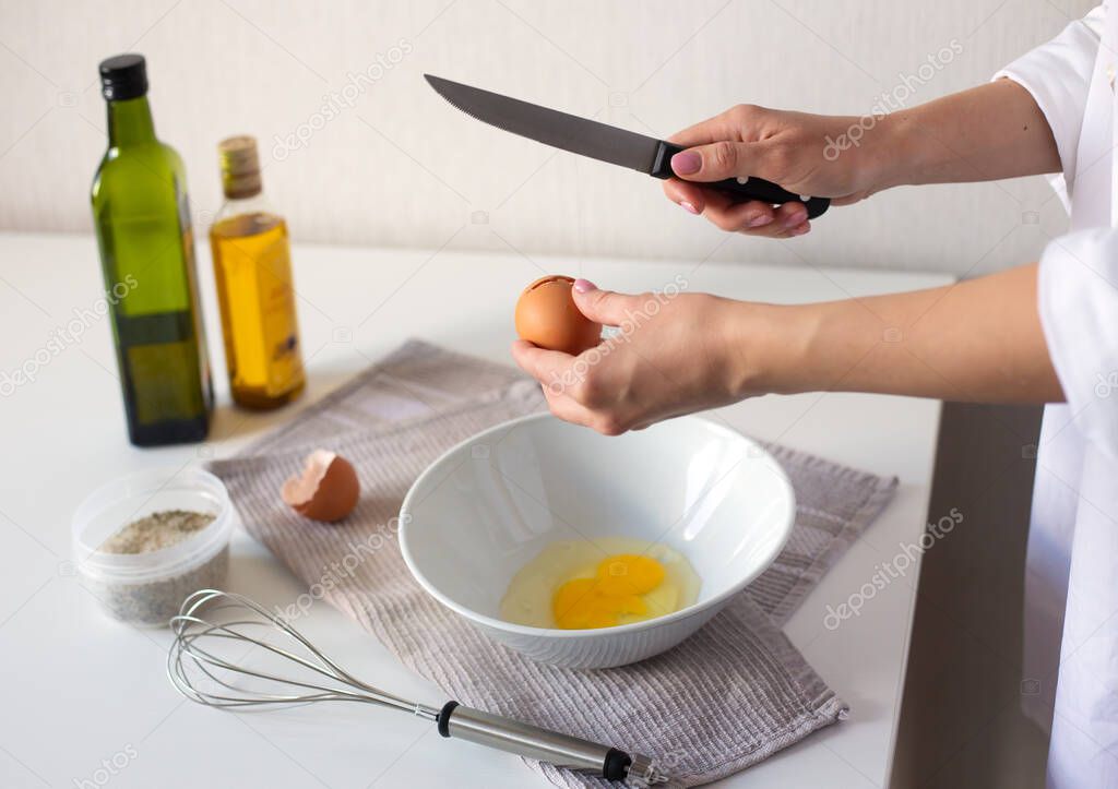 Female hands crack egg into bowl cooking omelet. Steel whisk, oil and vinegar bottles, salt and napkin on white table.
