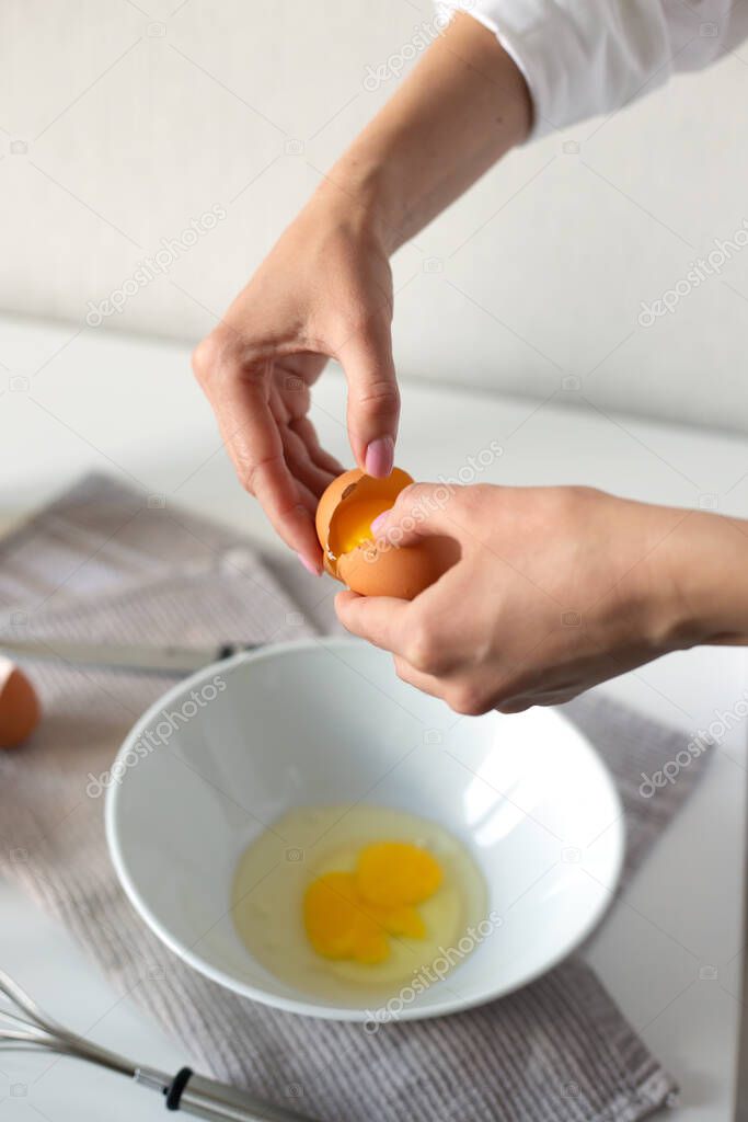 Female hands crack egg into bowl cooking omelet. Steel whisk, oil and vinegar bottles, salt and napkin on white table. Vertical