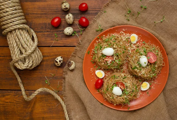 Bıldırcın yumurtalı, domatesli, mikro yeşil ve kahverengi ipli karabuğday eriştesi. Seçici odaklanma. Peynirli, sebzeli ve kırmızı tabakta roka kaplı karabuğday makarnası ahşap arka planda malzemelerle. Yatay, üst görünüm.