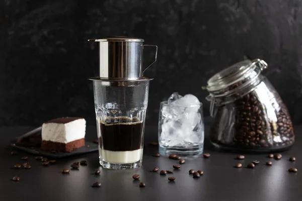Traditional vietnamese coffee maker placed on the top of glass, glass jar with coffee beans, glass with ice and souffle dessert. Iced coffee for summer. Coffee dripping into the glass with condensed milk at the bottom on grunge dark background. Low k