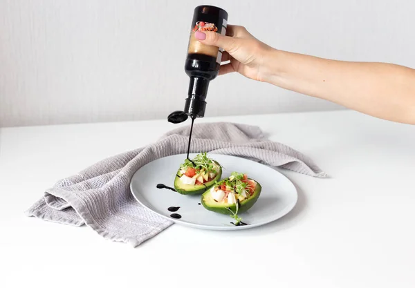 Avokado kasesine salatanın üzerine balzamik sosu döküyorum. Avokado, domates, mikroyeşillik, vejetaryen aperatifi beyaz arka planda gri peçeteyle beyaz tabakta sağlıklı vejetaryen yemeği.
