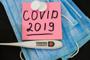 Koronavirüs enfeksiyonuyla mücadele sırasında yüksek ateş kavramı. Tıbbi maskelerin, termometrenin, tabletlerin arka planında COVID-2019 yazılıydı..