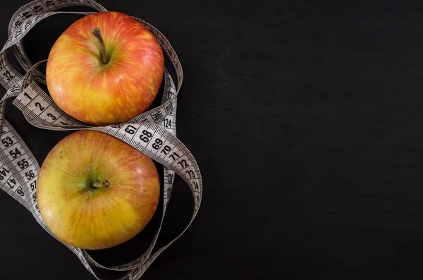 The concept of weight loss and diet. Two apples and a measuring tape on a black background. View from above. Copy space. Place for text.