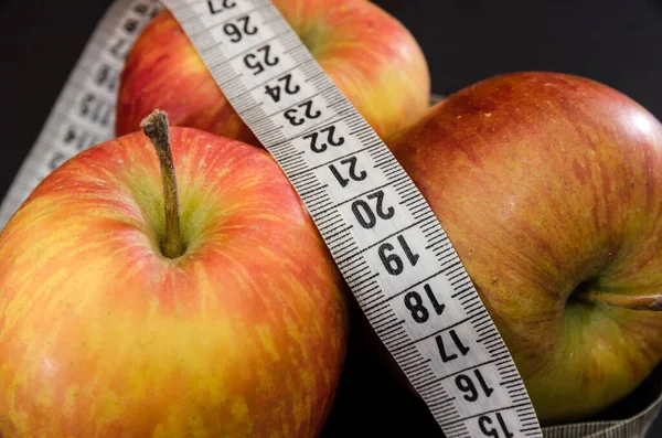 The concept of weight loss and diet. Three apples and a measuring tape on a black background. Close-up.