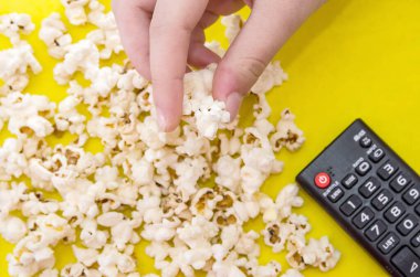 Patlamış mısır ve uzaktan kumanda, sarı arka planda üst görüntü. İnsanlar patlamış mısır yiyor. İnsan eli. Yukarıdan görüntüle.