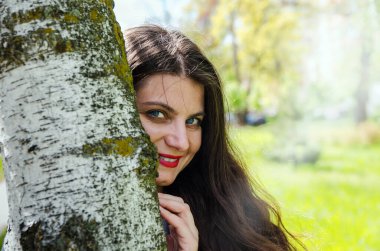 Güzel genç kız bir ağacın arkasından dışarı bakıyor. Yakın plan plan. Güneş ışığı, parkta yaz günü..