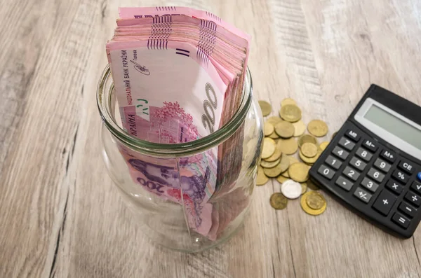 Cam bir kavanozda 200 Hryvnia, bozuk paralar ve ahşap bir arka planda hesap makinesi. Tasarruf kavramı.