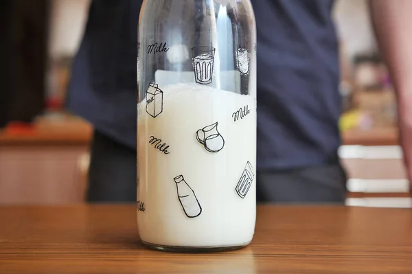 Egy üveg tej van az asztalon, egy friss tejet öntő személy hátterében. — Stock Fotó
