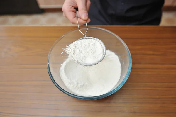 Peneirar farinha de trigo sobre uma tigela de vidro transparente na cozinha — Fotografia de Stock