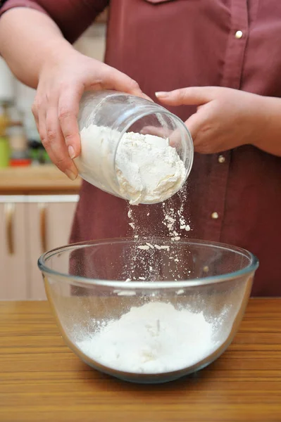 Dłonie kobiety wlewają mąkę pszenną do szklanej miski na stole. — Zdjęcie stockowe