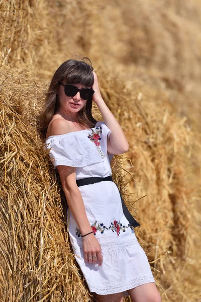 白いドレスと眼鏡のブルネットの女の子は 丸い玉からの大きな干し草の山の背景を背景に — ストック写真