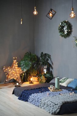 Gri duvara karşı tavan dekoru. Ekoseli alçak yatak, ahşap raf, sandalye, çelenkler ve eski Edison lambaları..