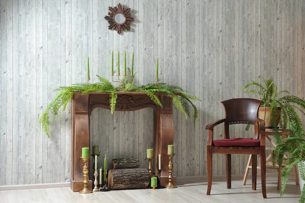 Interior with wood Wallpaper, wood fireplace, chair and fern, candles, copper candlesticks and small round mirror on the wall.