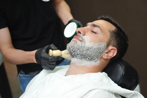 Hairdresser applying foam with brush on male face to shave beard.