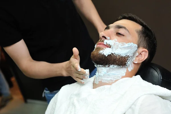 Hairdresser applying foam on male face to shave beard.