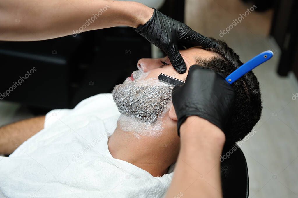 Hairdresser shaving male beard with blade in male barber shop. 