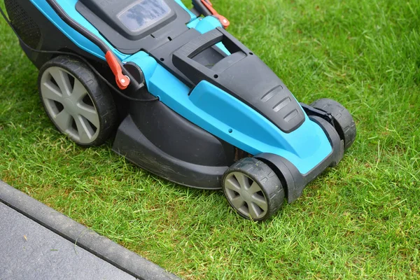 An electric lawn mower cuts the lawn near the edge and concrete curb.