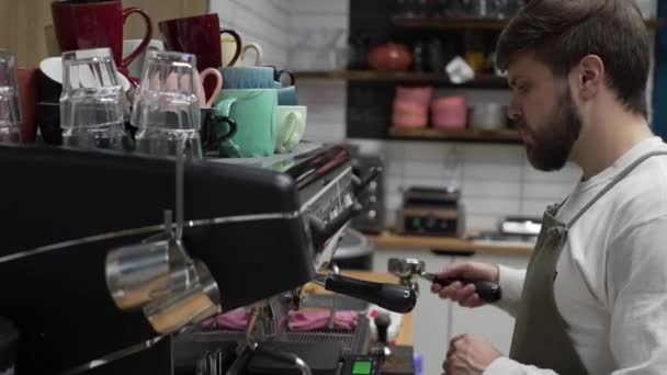 Sakallı bir erkek barista kahve makinesinin yanında duruyor ve ciddi bir bakışla kahve yapıyor. — Stok video