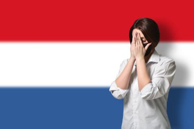 Stresli ve koruyucu maskeli bir kadın ellerini Hollanda bayrağının arka planına koyup yüzünü kapatıyor. Anksiyete ve korku kavramı, koronavirüs salgını