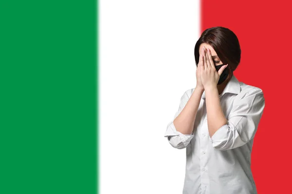 A woman in stress and a protective mask covers her face with her hands on the background of the flag of Italy. The concept of anxiety and fear, coronavirus pandemic — Stock Photo, Image